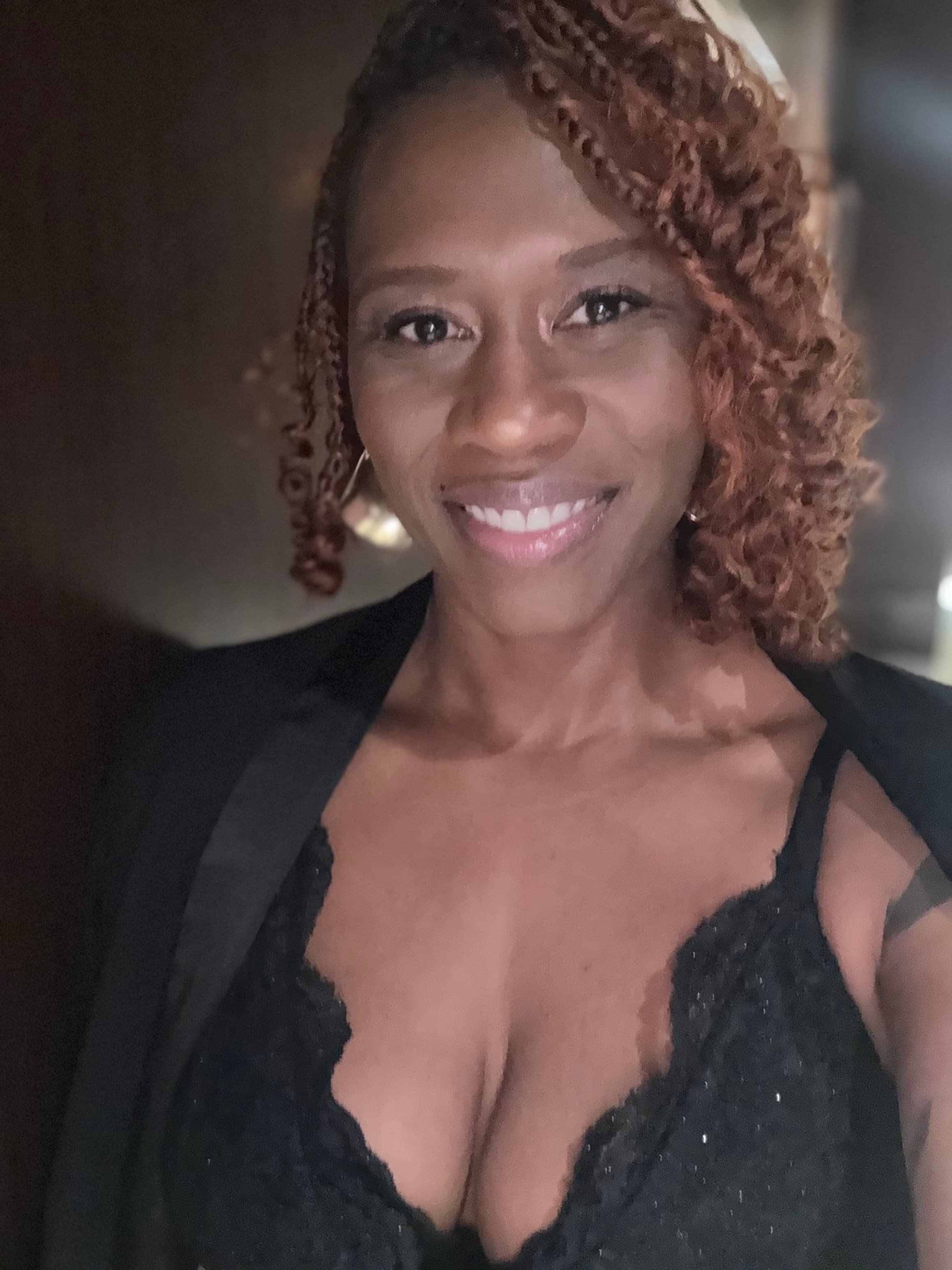 Smiling Black woman with auburn curls, wearing a black lace top and draped black blazer.